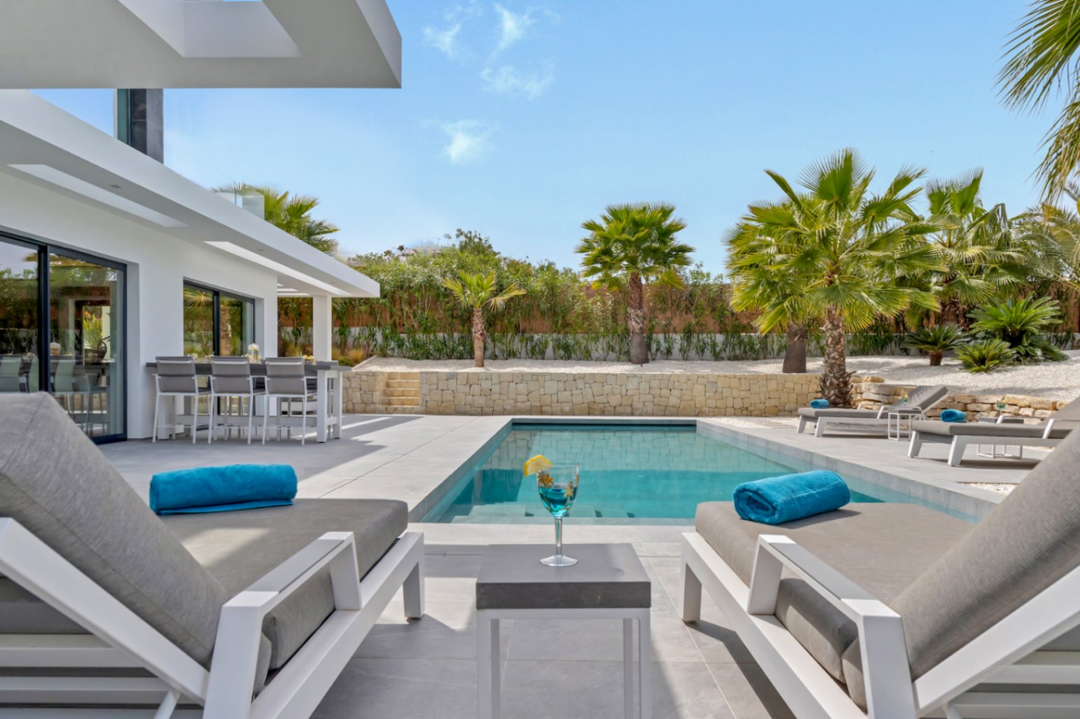 La moderna zona exterior presenta una piscina rectangular, cómodas tumbonas y una barra integrada con taburetes altos, todo ello enmarcado por un exuberante paisaje de palmeras.