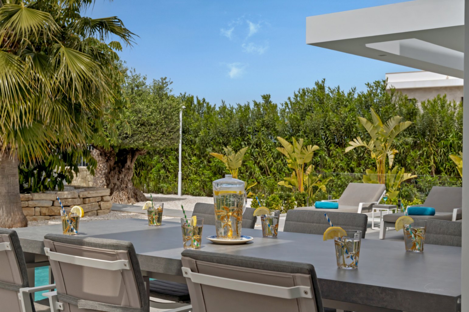 La zona de comedor exterior presenta una mesa gris moderna con bebidas refrescantes, rodeada de exuberante vegetación mediterránea, tumbonas y una vista parcial de la piscina.