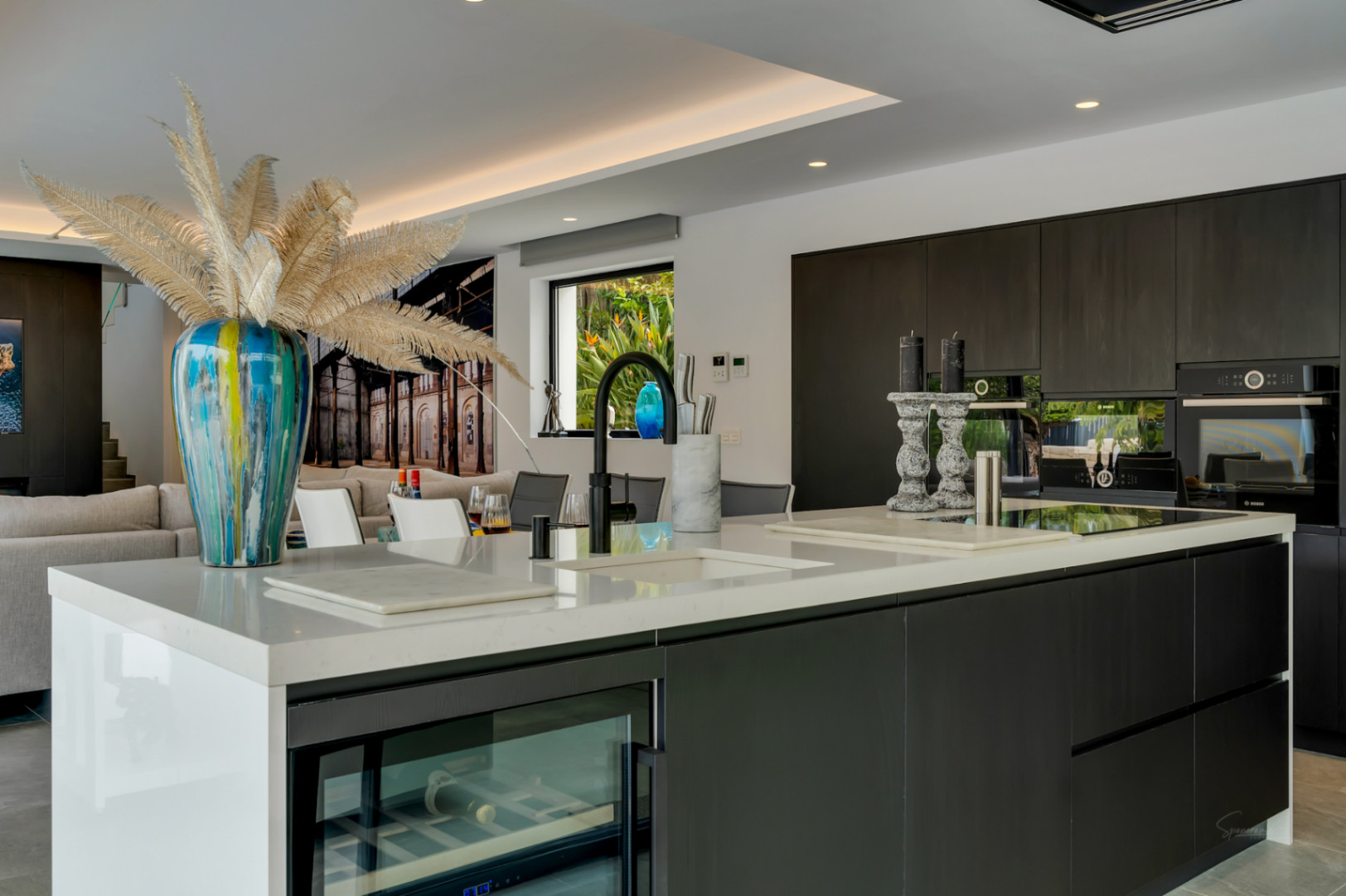 Moderna cocina de concepto abierto con isla de cuarzo blanco, gabinetes oscuros con electrodomésticos integrados y vinoteca, con vistas a un jardín exuberante.