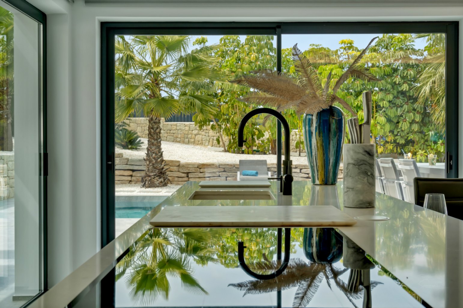Moderna isla de cocina con grifo negro y fregadero integrado, ofreciendo una vista panorámica al jardín mediterráneo con palmeras, muros de piedra y la piscina a través de amplias puertas de cristal.
