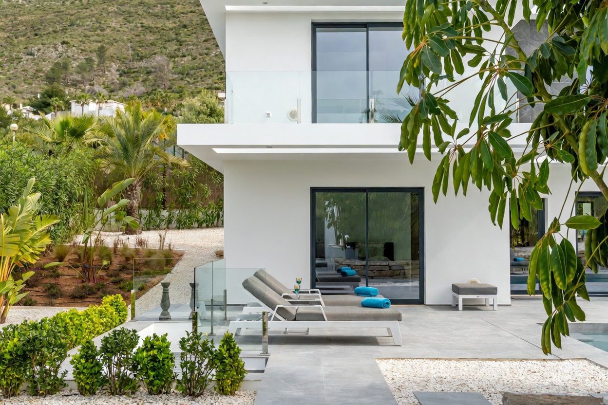 Moderna terraza exterior con suelo de baldosas grises y tumbonas contemporáneas, delimitada por una barandilla de cristal con vistas a un exuberante jardín mediterráneo y montañas lejanas.