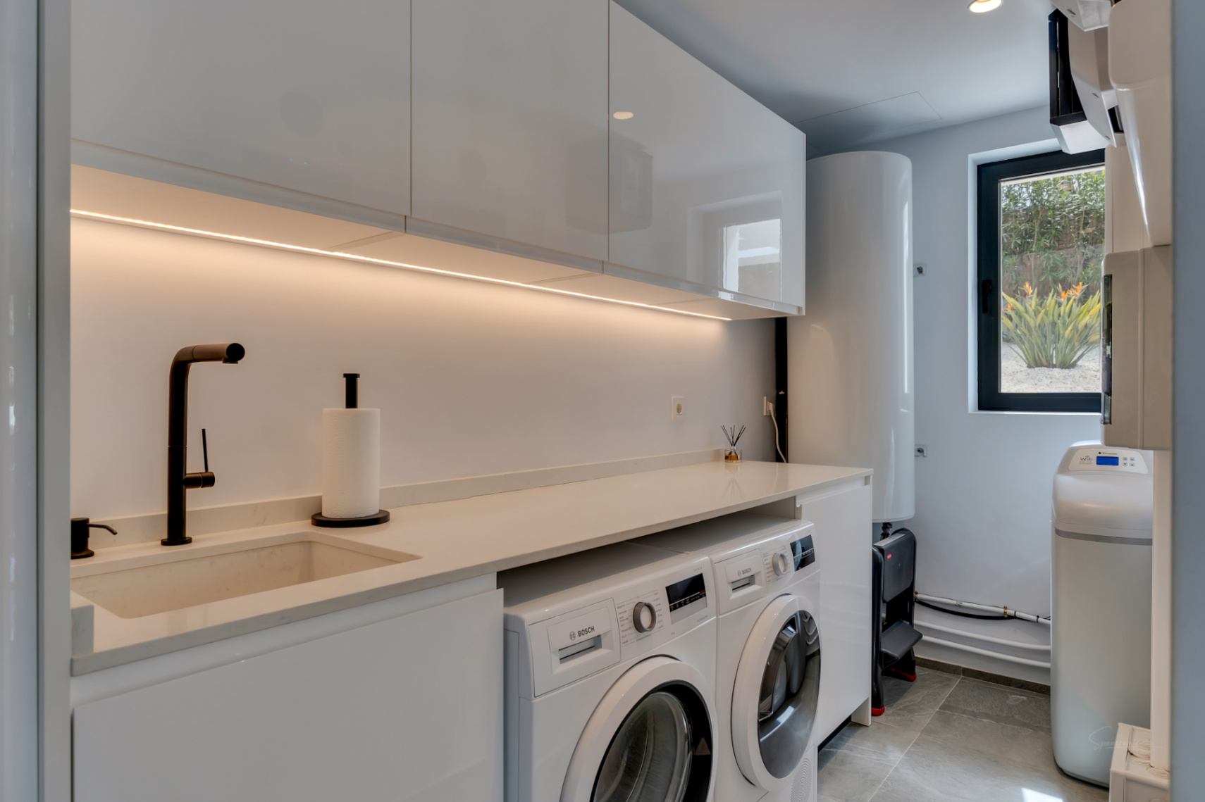Moderna zona de lavandería con gabinetes blancos lacados, fregadero integrado, grifo negro mate y electrodomésticos Bosch, realzada por iluminación LED bajo mueble y vista a la vegetación.