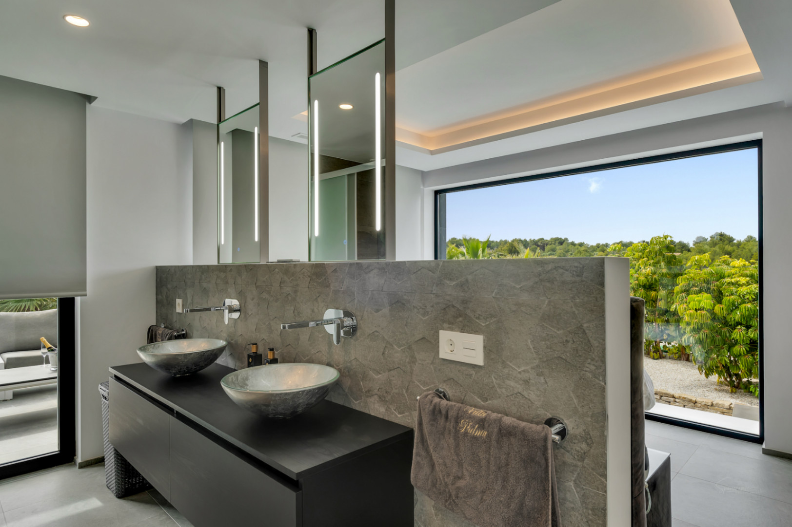 Moderno baño con doble lavabo, lavamanos de diseño y grifería mural. Espejos iluminados y una gran ventana ofrecen vistas a la exuberante vegetación.