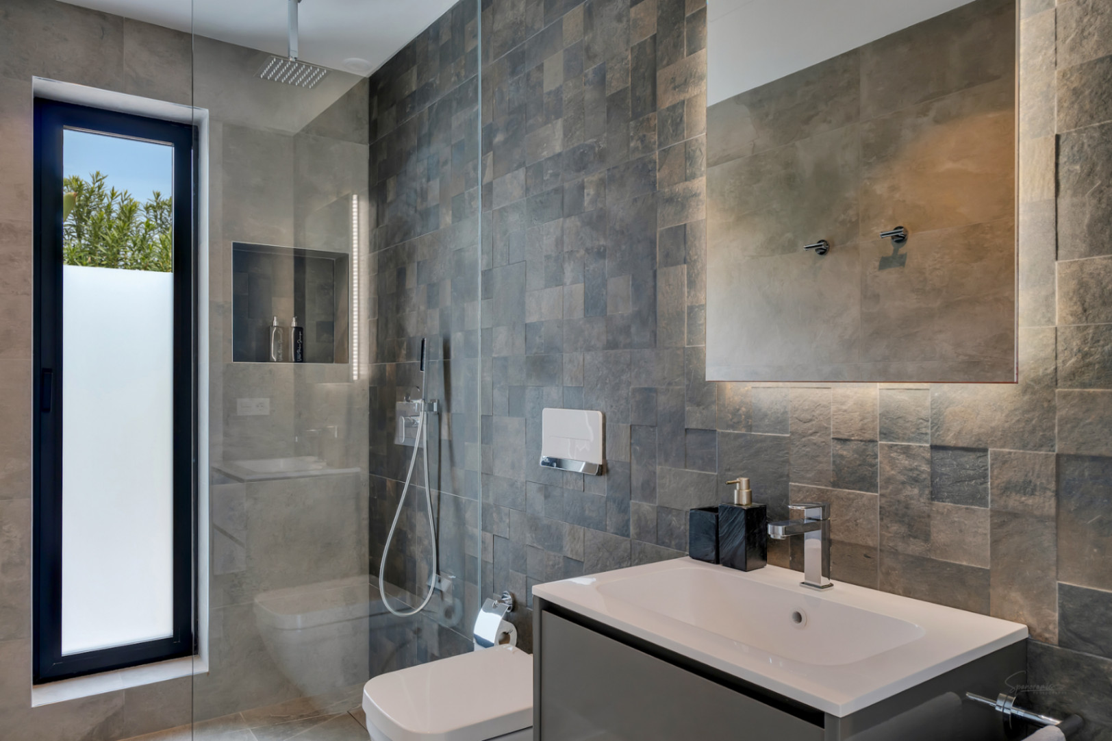 Moderno baño con ducha de lluvia y mampara de cristal. Paredes de azulejos texturizados, ventana esmerilada y tocador con lavabo integrado. Espejo retroiluminado.