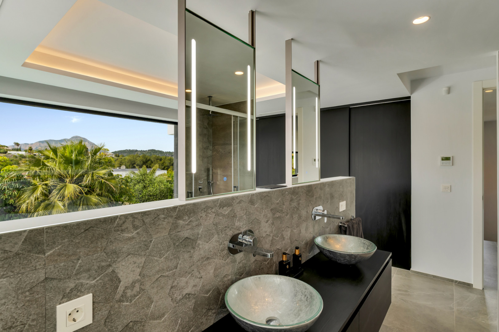 Moderno baño con lavabos dobles de cristal, salpicadero de azulejos hexagonales grises texturizados y espejos suspendidos iluminados. Amplia ventana con vistas a la montaña.