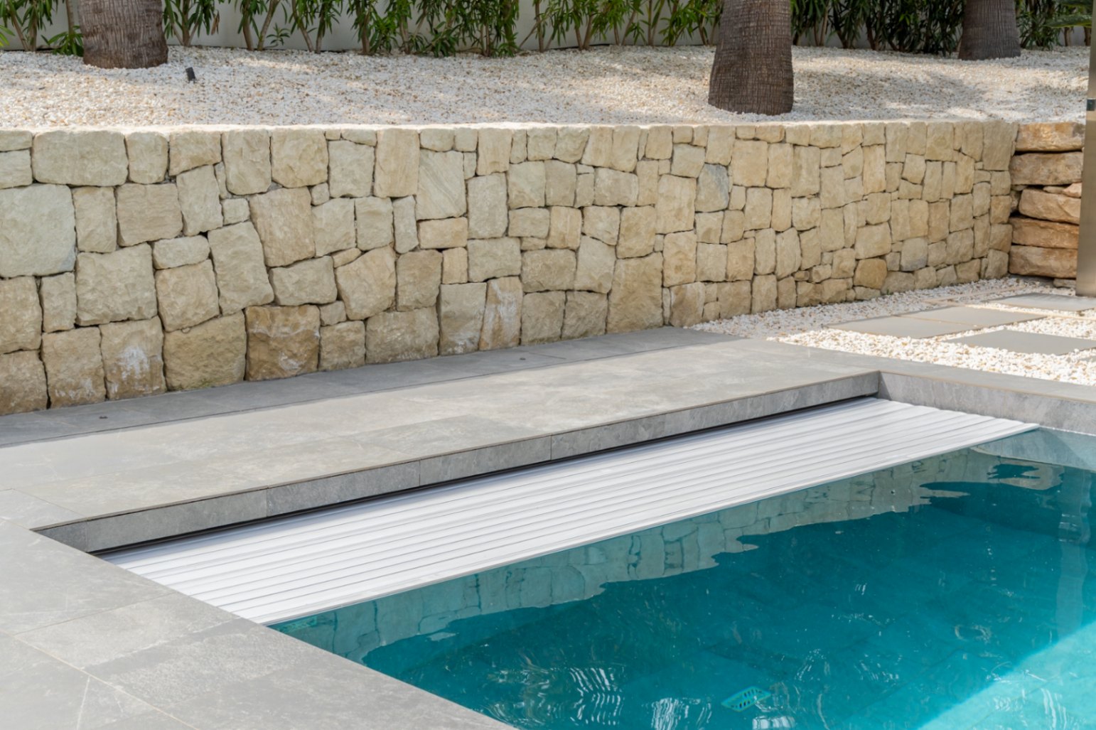 Piscina con cubierta de lamas integrada y borde de gres gris. Muro de piedra natural, jardín con grava blanca y palmeras al fondo.