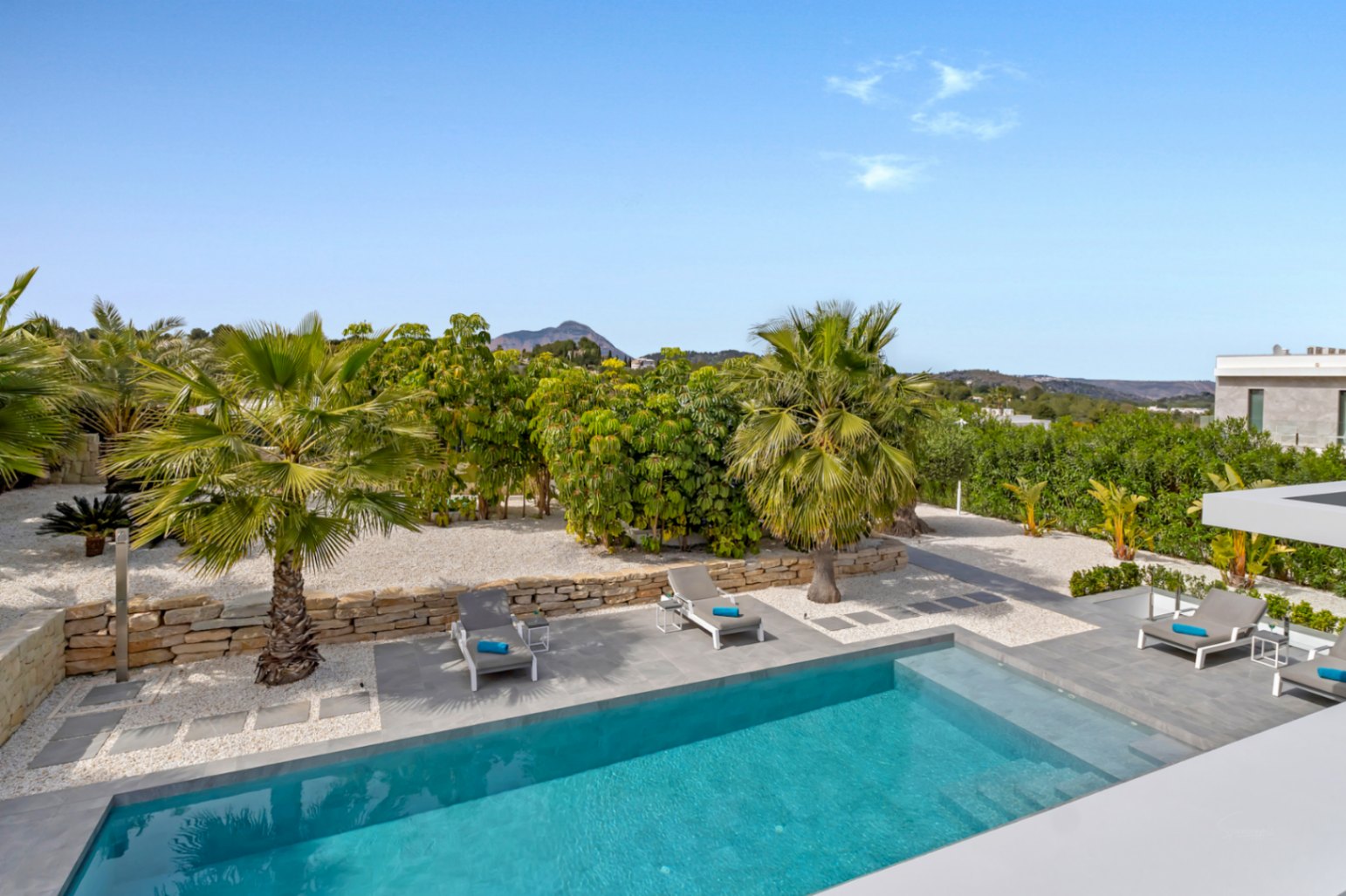Piscina rectangular de agua azul rodeada por una terraza de gres gris, tumbonas y un jardín mediterráneo con palmeras, muro de piedra y vistas a la montaña.