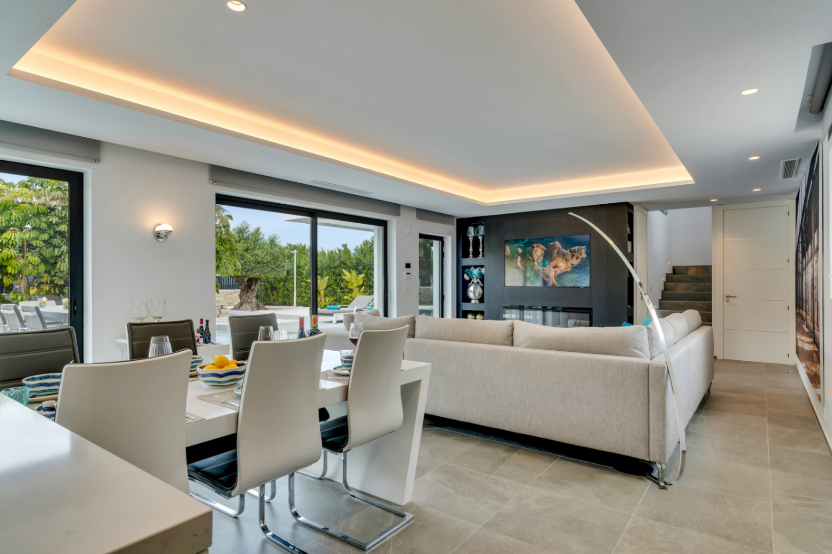 Salón-comedor de planta abierta con mesa blanca moderna, sofá gris claro y pared multimedia con chimenea eléctrica. Grandes puertas de cristal conectan con el jardín.
