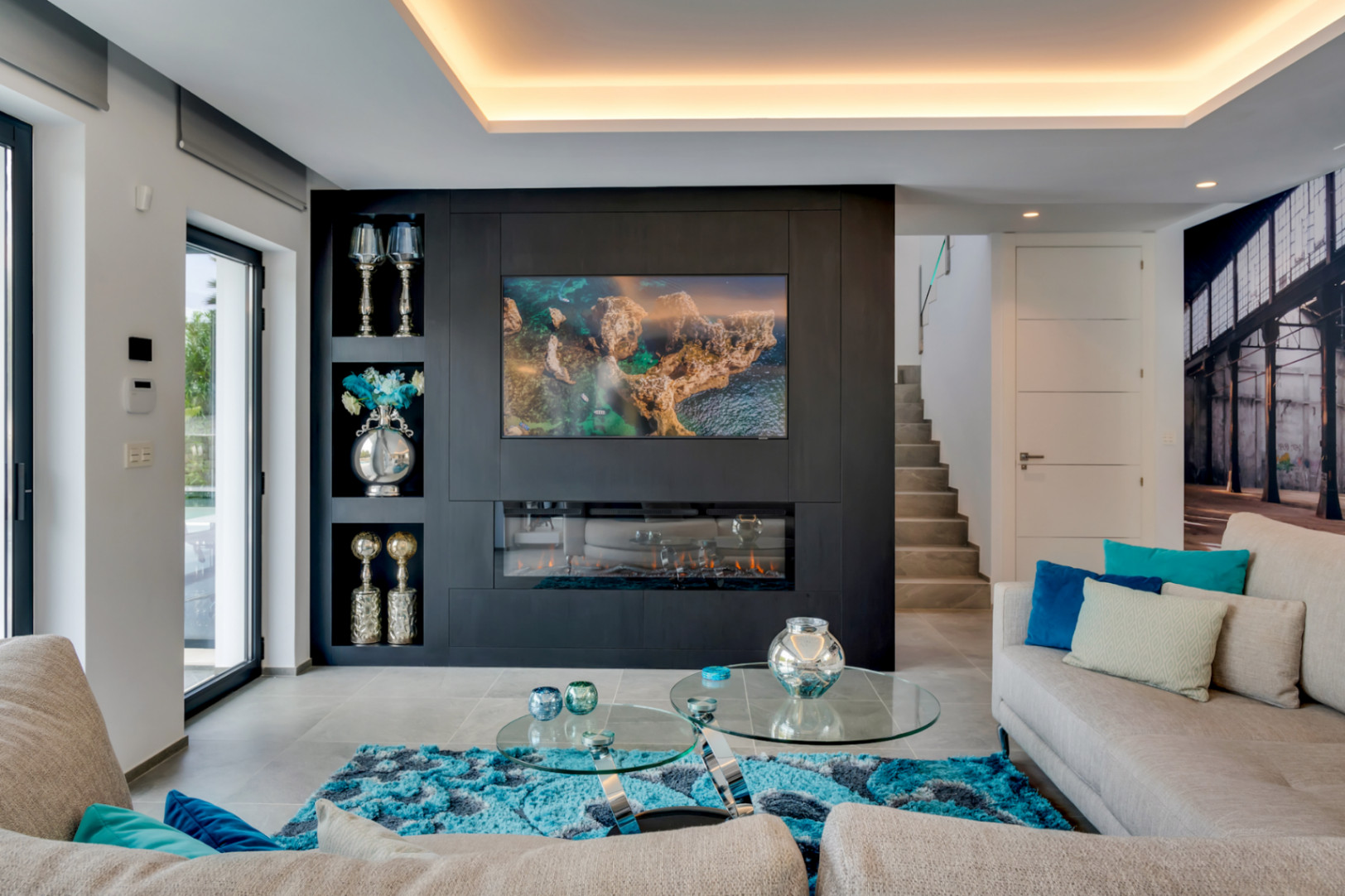 Salón moderno con pared decorativa oscura, TV integrada y chimenea eléctrica. Grandes puertas de cristal aportan luz natural y acceso al exterior, complementado por iluminación de techo.