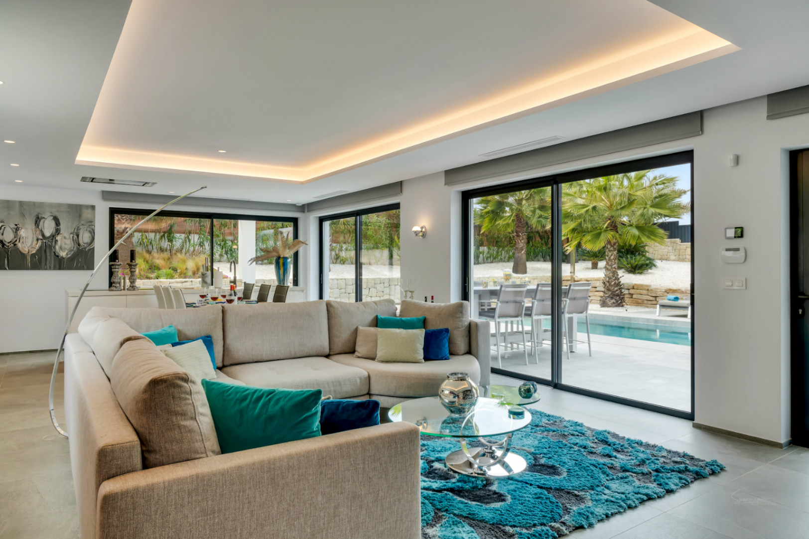 Salón moderno con sofá en L, alfombra turquesa y amplios ventanales que ofrecen acceso directo a la piscina y vistas a las palmeras.