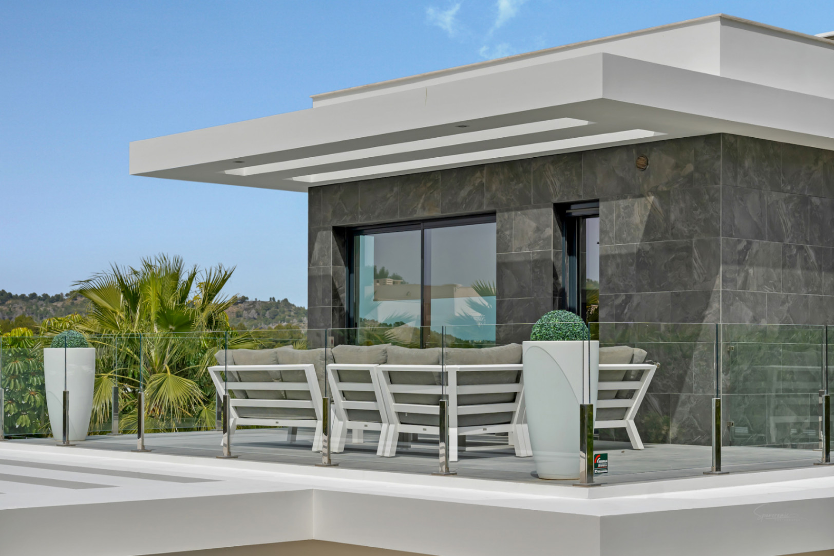 Terraza exterior con mobiliario moderno de diseño, barandilla de cristal y fachada de azulejos grises. Vistas a la vegetación y colinas.