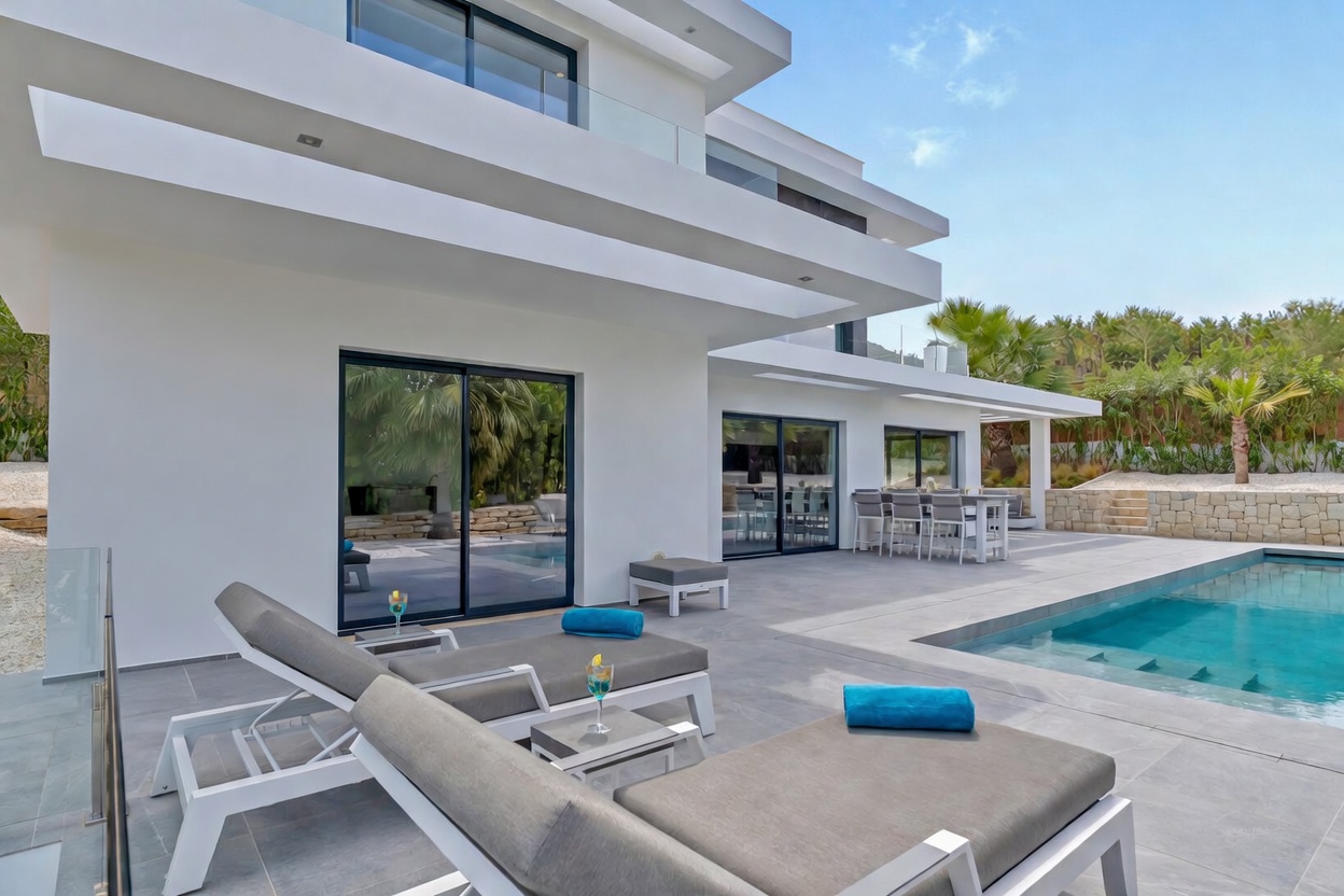 Terraza exterior con piscina de diseño rectangular, rodeada de tumbonas modernas y una zona de comedor al aire libre, conectada a la villa por amplios ventanales.