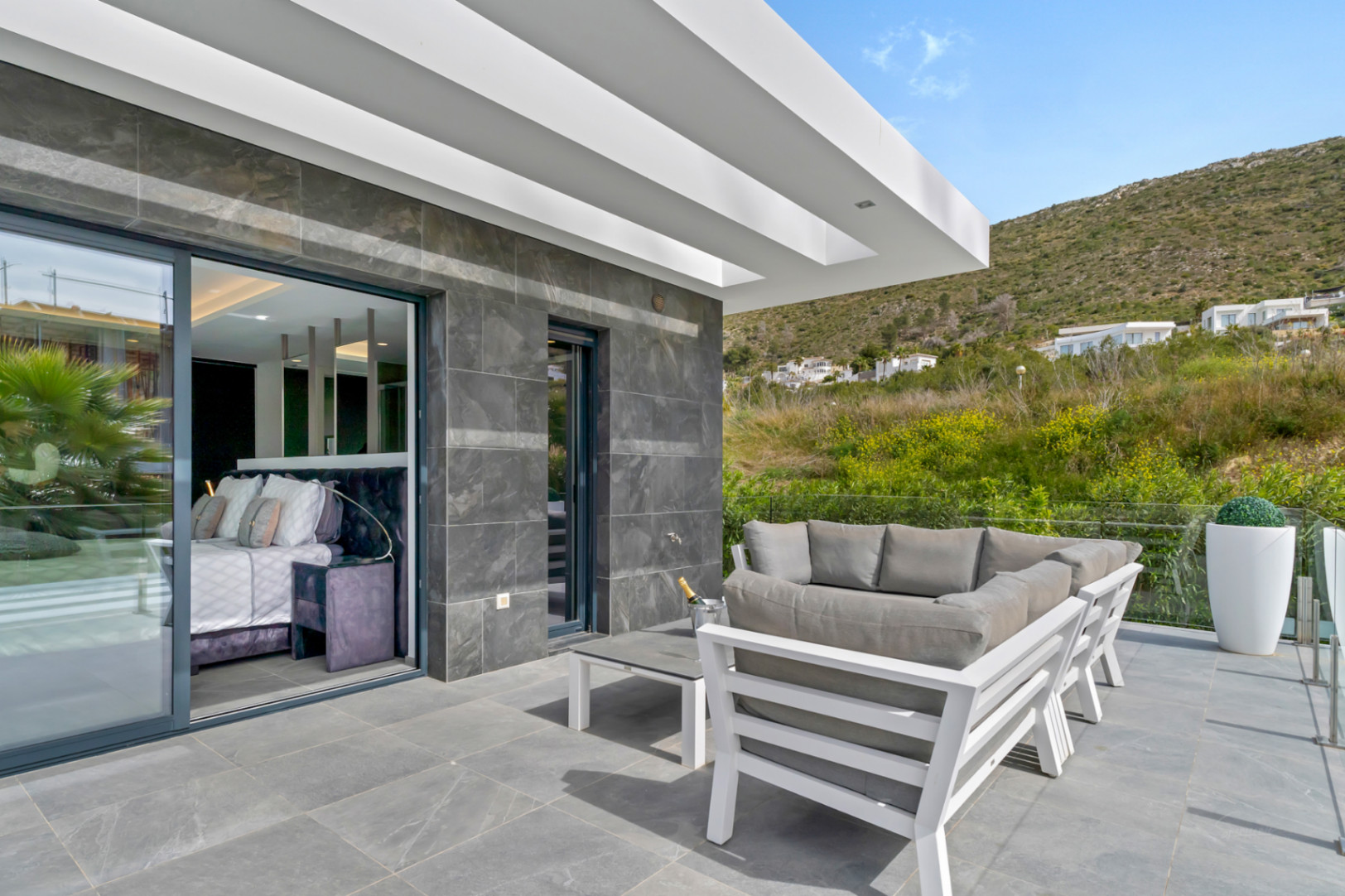 Terraza exterior con suelo de gres gris y elegante mobiliario de salón blanco. Grandes puertas correderas conectan con un dormitorio interior, ofreciendo vistas a la colina verde.