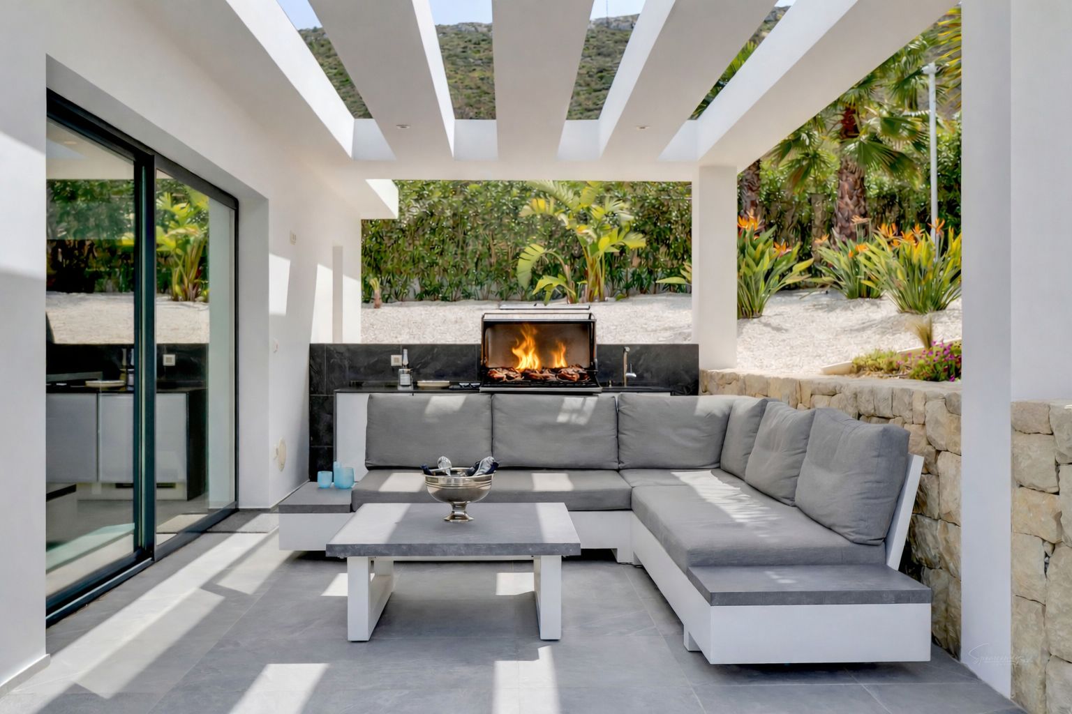 Terraza exterior cubierta con cocina integrada y barbacoa encendida, equipada con un sofá en L y mesa de centro sobre suelo de gres gris, frente a un jardín paisajístico.