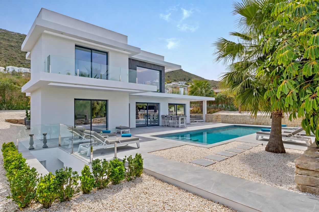 Villa blanca moderna con amplios balcones de cristal y piscina rectangular. El exterior cuenta con patio de baldosas grises, paisajismo de grava, comedor exterior y tumbonas, con fondo montañoso.