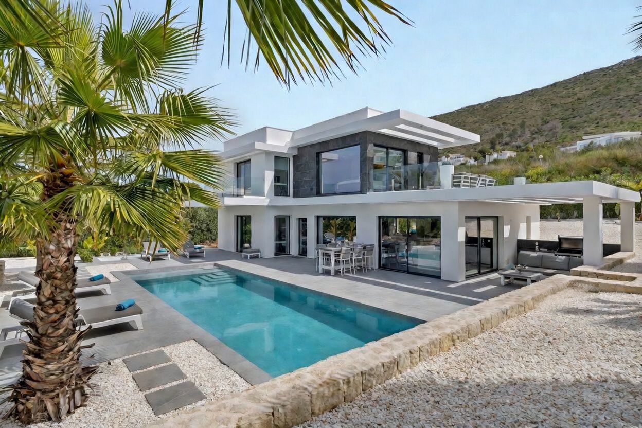 Vista exterior de la villa moderna, con su piscina rectangular, amplia terraza pavimentada con tumbonas y dos áreas de estar exteriores cubiertas, enmarcada por una palmera y una ladera natural.
