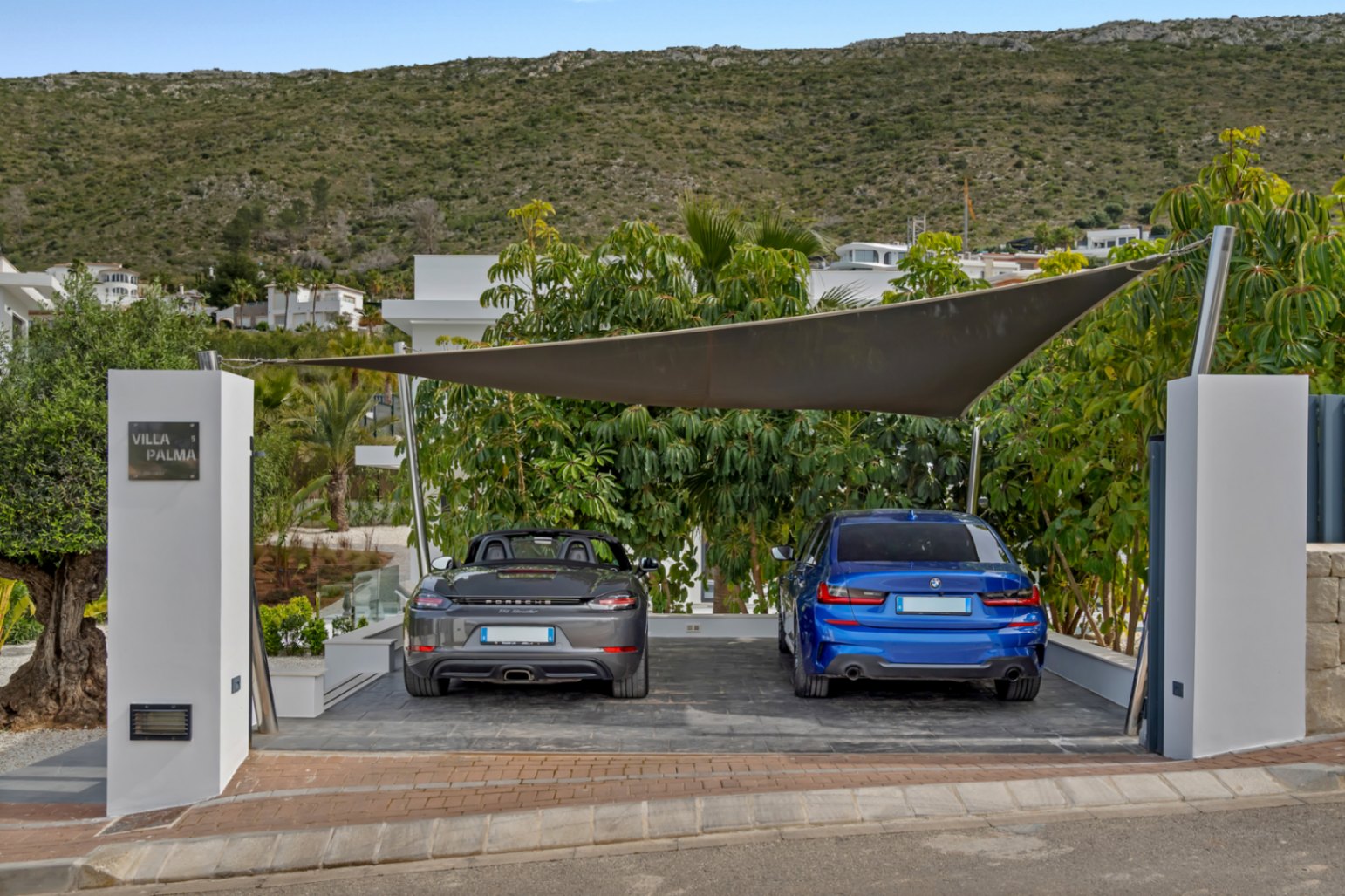 Zona de aparcamiento exterior con vela de sombra moderna, para dos coches de lujo: un Porsche gris y un BMW azul. El pilar de entrada muestra el letrero 'Villa Palma', rodeado de vegetación mediterránea y una colina al fondo.