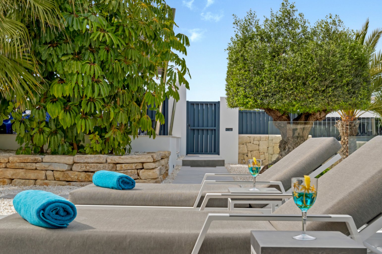 Zona de relax exterior con dos tumbonas modernas, toallas azules y cócteles. Un sendero conduce a un portón azul, flanqueado por exuberante vegetación y un muro de piedra.
