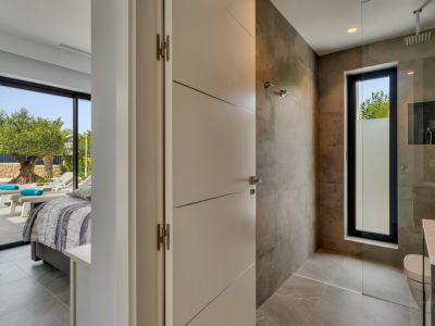 Acceso a un moderno baño con ducha de obra y ventana esmerilada, junto a un dormitorio con salida directa a terraza y vistas a un olivo. Suelos y paredes de gres en tonos neutros.