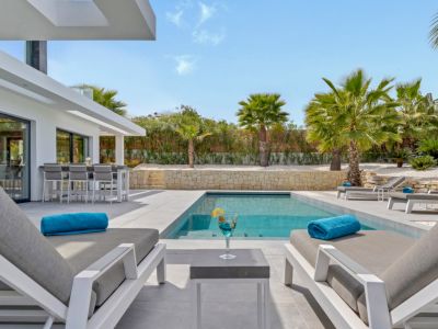 Amplia terraza con piscina rectangular de diseño, elegantes tumbonas y barra exterior integrada, conectada a la vivienda y rodeada de palmeras.