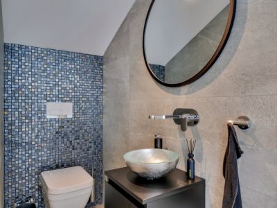Aseo moderno con pared de mosaico en tonos azules y dorados, lavabo de diseño y espejo redondo grande. Azulejos de gran formato en gris complementan el elegante diseño.