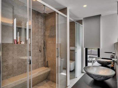 Baño moderno con amplia ducha de obra, revestida en piedra texturizada y equipada con banco integrado. El tocador cuenta con dos lavabos de cristal sobre encimera oscura, con vistas a una zona exterior.