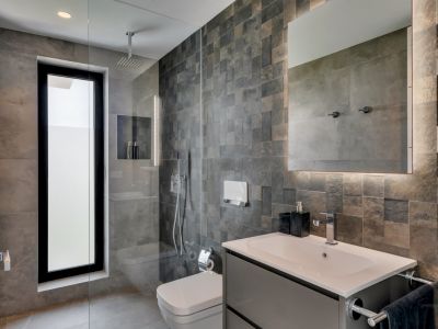 Baño moderno con ducha de obra, ventana vertical y azulejos grises texturizados. Lavabo flotante con espejo retroiluminado y sanitarios contemporáneos.