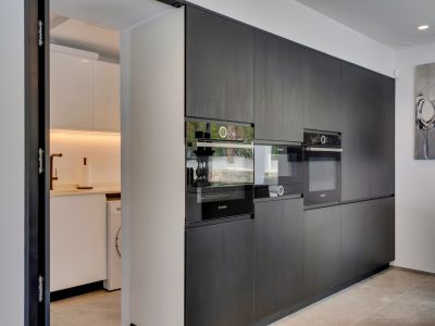 Cocina de concepto abierto con elegantes armarios oscuros integrados y electrodomésticos Bosch, conectada discretamente a una despensa funcional con lavadora y grifo cobrizo.