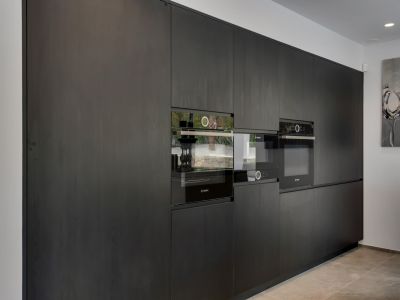 Cocina moderna con mobiliario oscuro de madera de suelo a techo y electrodomésticos Bosch integrados. Incluye horno y microondas encastrados, con reflejos del exterior.