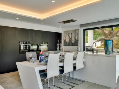 Cocina moderna de concepto abierto con isla-comedor integrada, armarios oscuros y hornos empotrados, conectada a un jardín a través de grandes ventanales.