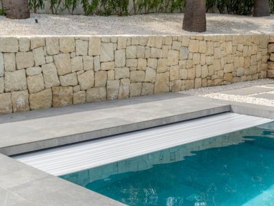 Cubierta automática de lamas blancas protegiendo la piscina, rodeada de pavimento gris y un muro de piedra natural, con jardín de grava blanca al fondo.
