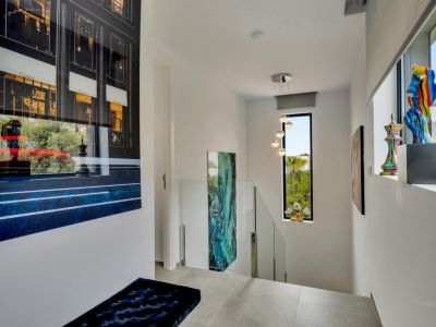 Distribuidor de escalera moderno con barandillas de cristal y suelo de gres. Gran obra de arte de Cartier, ventana vertical con vistas a la vegetación y lámpara colgante esférica. Esculturas coloridas en el alféizar.