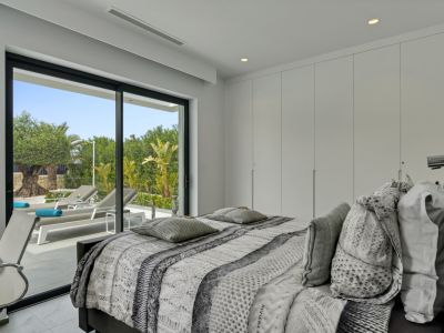 Dormitorio moderno con acceso directo a una terraza privada. Grandes puertas correderas de cristal muestran tumbonas y vegetación mediterránea, complementado por armarios empotrados.
