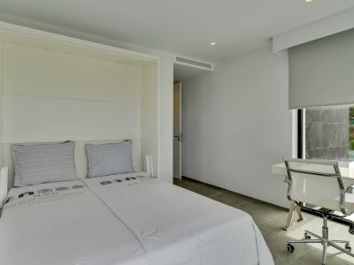 Dormitorio moderno con cama integrada y zona de estudio junto a una ventana con vistas parciales a una terraza exterior y vegetación. Suelo de gres gris y paredes blancas.