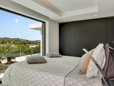Dormitorio moderno con ventanal de suelo a techo que enmarca vistas panorámicas a colinas verdes. Armarios empotrados oscuros y elegante iluminación empotrada completan el diseño.