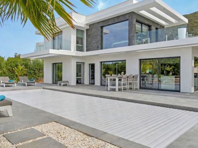 El exterior de la villa moderna presenta una piscina cubierta y una amplia terraza de azulejos con comedor al aire libre y tumbonas. Grandes ventanales y balcones con barandillas de cristal.
