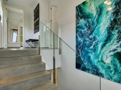 Escalera de piedra con barandilla de cristal, destacando una gran obra de arte abstracta azul-verde. Arriba, un pasillo con espejo decorativo y banco.