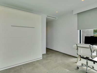 Espacio multifuncional con cama abatible integrada, escritorio moderno frente a una ventana con persiana y suelo de baldosas grises.