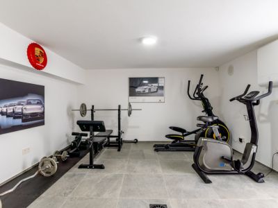 Espaciosa sala de fitness equipada con banco de pesas, barra, mancuernas, elíptica y bicicleta estática. Presenta paredes blancas, suelo de gres gris y decoración automovilística.