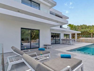 Exterior de villa moderna con piscina de diseño, zona de solárium con tumbonas y comedor al aire libre, integrado en un paisaje mediterráneo.
