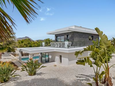 Exterior de villa moderna de varios niveles con revestimiento blanco y piedra oscura. Destaca una terraza superior con barandillas de cristal y asientos, rodeada de grava blanca y plantas tropicales.