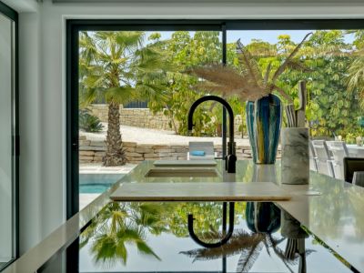 Isla de cocina moderna con superficie reflectante y grifo negro. Grandes puertas de cristal enmarcan vistas a palmeras, muros de piedra y una tranquila piscina, fusionando interior y exterior.