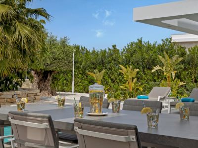 La zona de comedor exterior presenta una mesa gris moderna con bebidas refrescantes, rodeada de exuberante vegetación mediterránea, tumbonas y una vista parcial de la piscina.