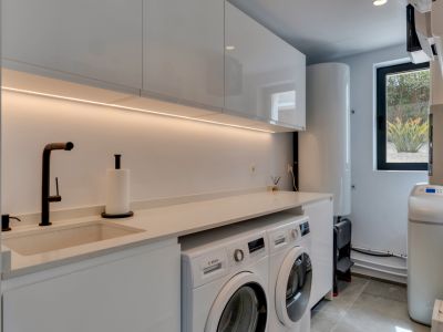 Lavadero moderno con armarios blancos de alto brillo, fregadero integrado y electrodomésticos Bosch. Iluminación LED bajo mueble y ventana con vista a la vegetación.