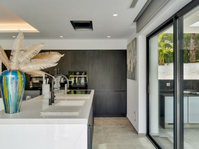 Moderna cocina de concepto abierto con isla central blanca, doble fregadero y grifería negra, conectada a un jardín de gravilla con cocina exterior a través de amplios ventanales.