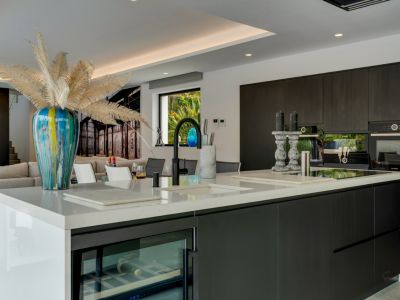 Moderna cocina de concepto abierto con isla de cuarzo blanco, gabinetes oscuros con electrodomésticos integrados y vinoteca, con vistas a un jardín exuberante.