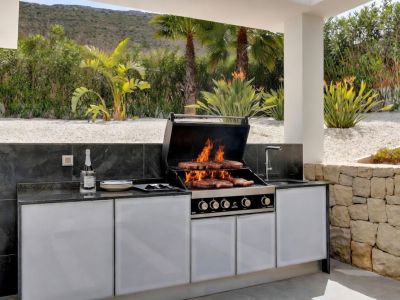 Moderna cocina exterior con barbacoa de gas integrada cocinando alimentos, encimeras de piedra oscura y gabinetes blancos, en un jardín paisajístico con grava y vegetación exuberante.