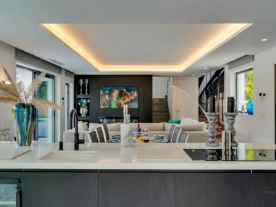 Moderna isla de cocina de concepto abierto con encimera de cuarzo blanco, fregadero integrado y placa de inducción, con vistas al comedor y sala de estar con pared de TV y luz de techo.