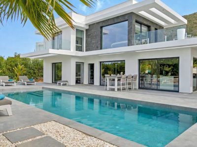 Moderna villa con piscina rectangular y amplia terraza de gres. Grandes ventanales conectan el interior, complementado por un comedor exterior y vistas a la montaña.