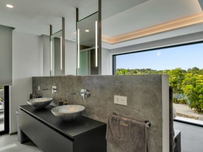 Moderno baño con doble lavabo, lavamanos de diseño y grifería mural. Espejos iluminados y una gran ventana ofrecen vistas a la exuberante vegetación.